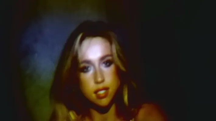 A stylized, dimly lit close-up of Tate Mcrae with long blonde hair, shown in soft focus with dramatic shadows and warm tones, creating a moody, music-video-like atmosphere