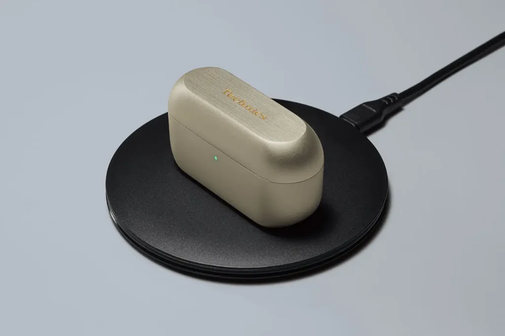 Technics EAH-AZ100 Champagne Gold charging case resting on a wireless charging pad, with a green indicator light illuminated, emphasizing its Qi-compatible design and brushed metallic finish