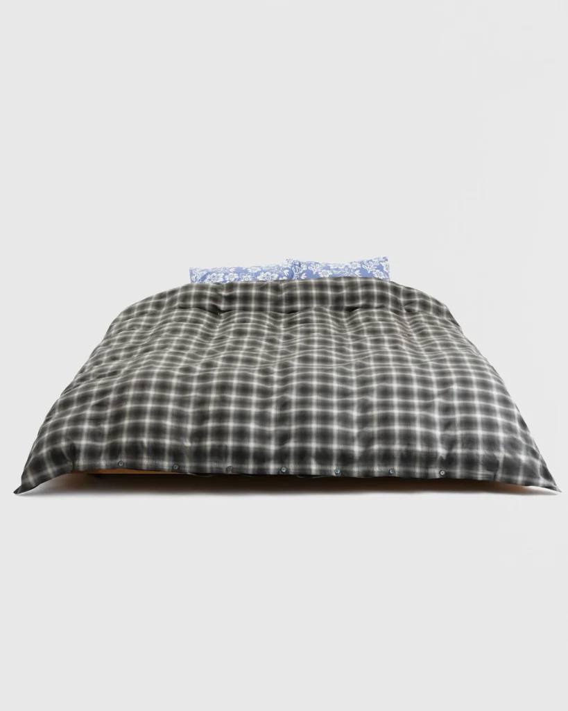 Soft black and grey plaid Stüssy x Tekla duvet cover on a neatly made bed, shown from the front with blue floral pillows peeking out behind it