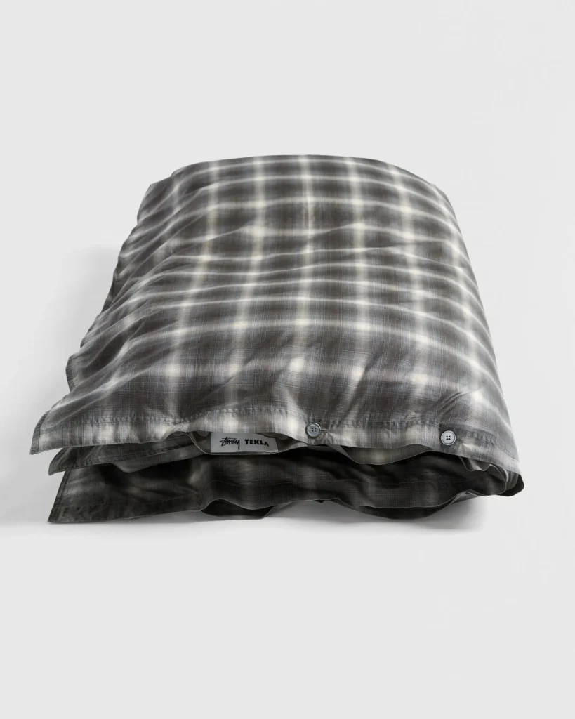 Folded Stüssy x Tekla Black Plaid duvet cover stacked neatly, showing the soft black-and-grey check pattern, button closure, and co-branded label peeking out.