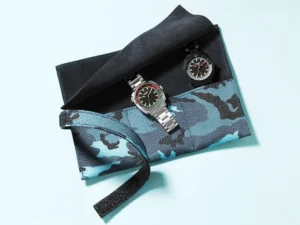 Aqua blue and petrol black camouflage three-pocket watch roll made of Cordura canvas, opened to reveal two wristwatches resting inside a soft black suede interior, displayed on a light blue background