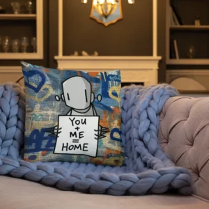 A decorative throw pillow featuring Chris RWK’s signature robot character holding a sign that reads “You + Me = Home,” set against a graffiti-inspired blue, orange, and beige collage background, placed on a plush light-purple knitted blanket draped over a beige tufted sofa in a cozy living room with a fireplace and shelves in the background