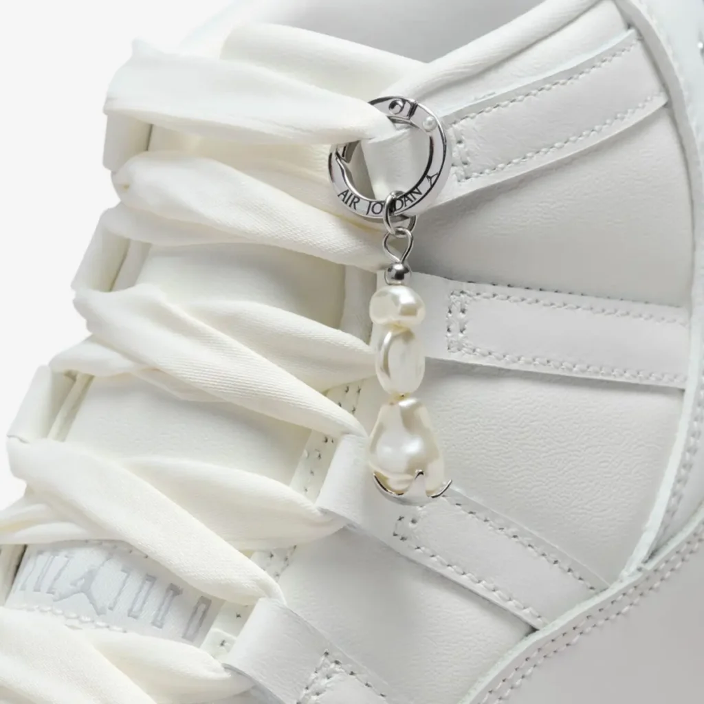 Close-up of the Air Jordan 11 WMNS “Pearl” detailing, showing silky white laces and a silver Jumpman-branded ring charm adorned with irregular pearl beads against the smooth white leather upper