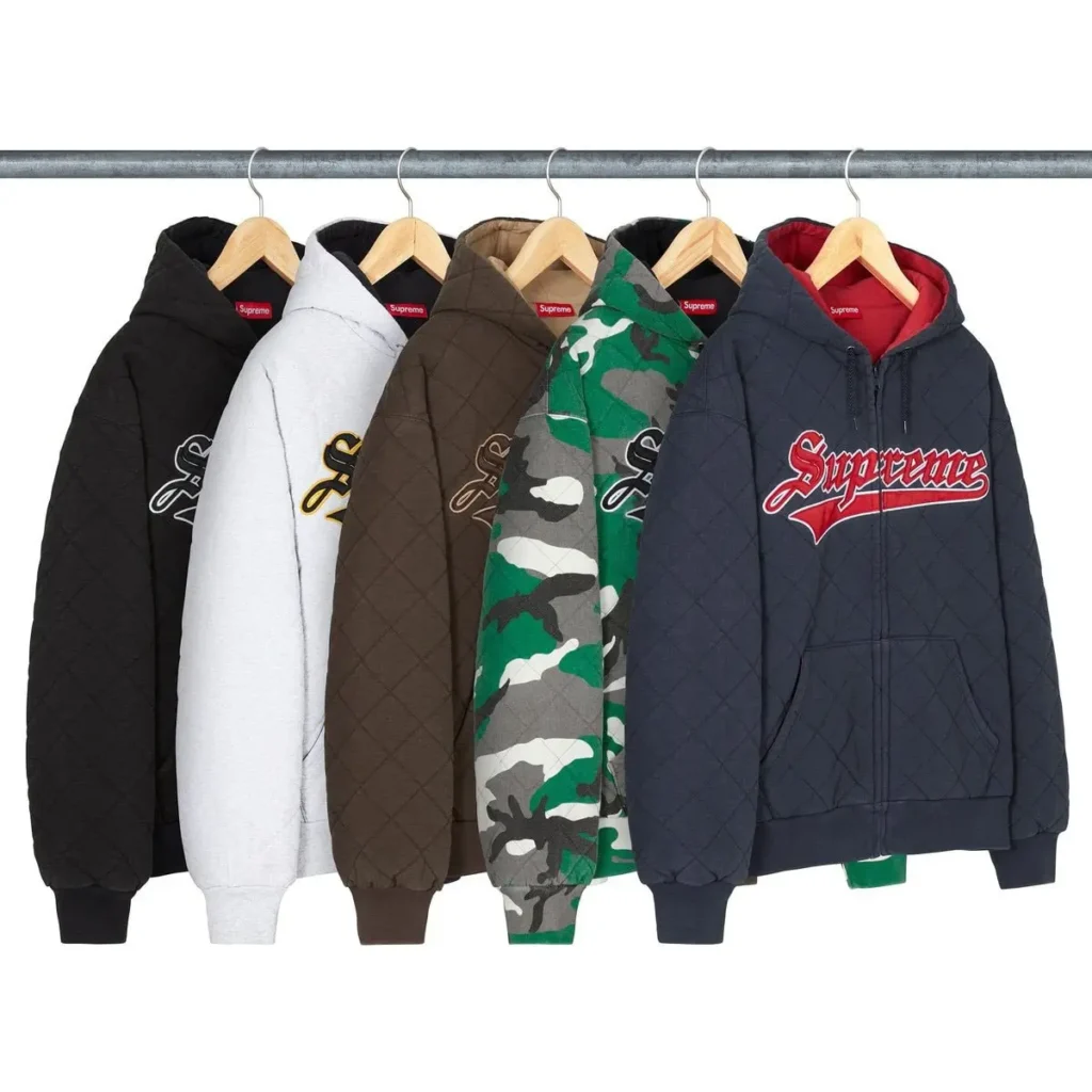A row of Supreme Fall/Winter 2025 quilted zip-up hoodies hanging on wooden hangers, displayed in black, grey, brown, green camo, and navy colorways, each featuring a stitched quilt pattern and embroidered Supreme script logo on the chest