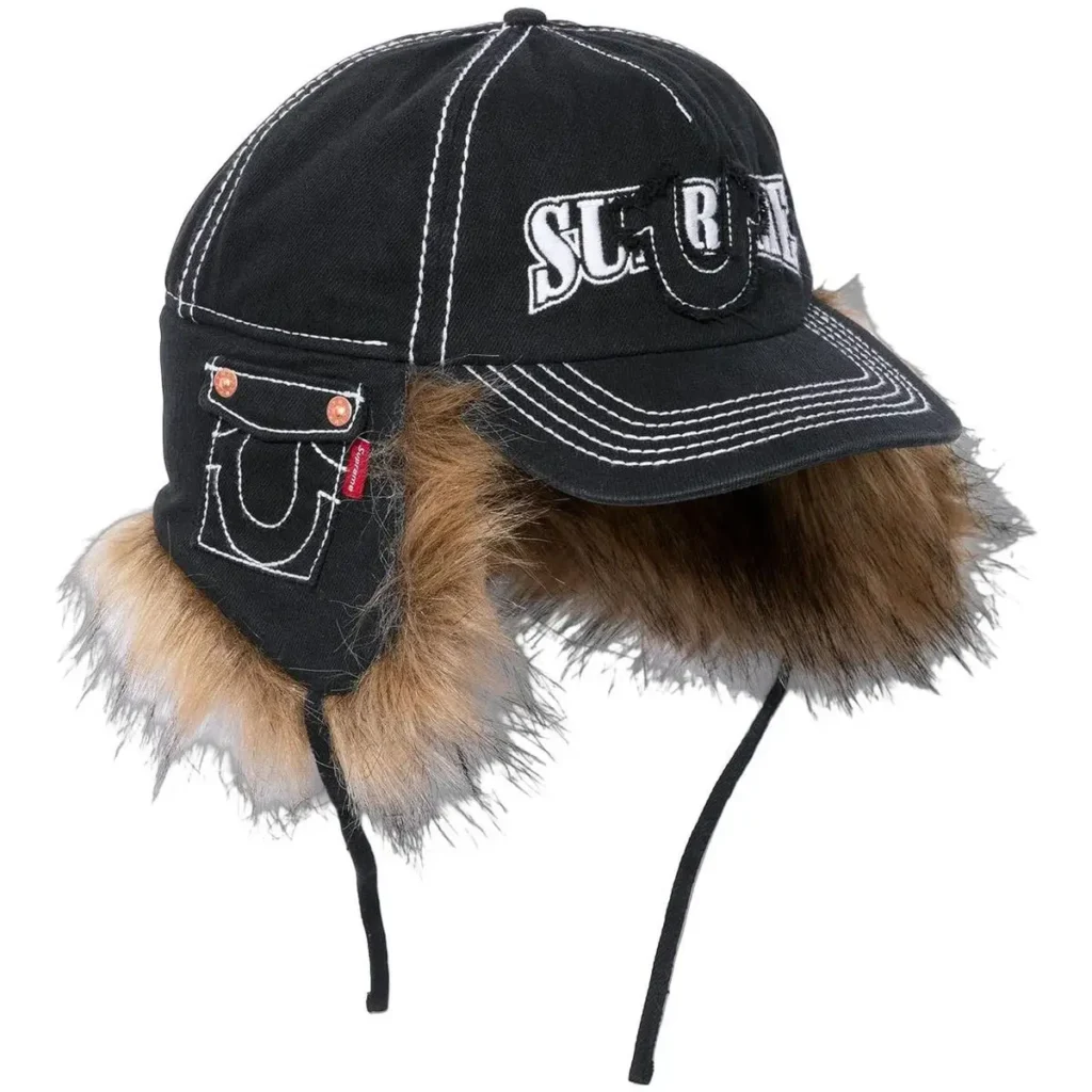 A black Supreme × True Religion earflap cap featuring contrast white stitching, a denim pocket with the True Religion horseshoe design on the side, and brown faux-fur lining around the earflaps