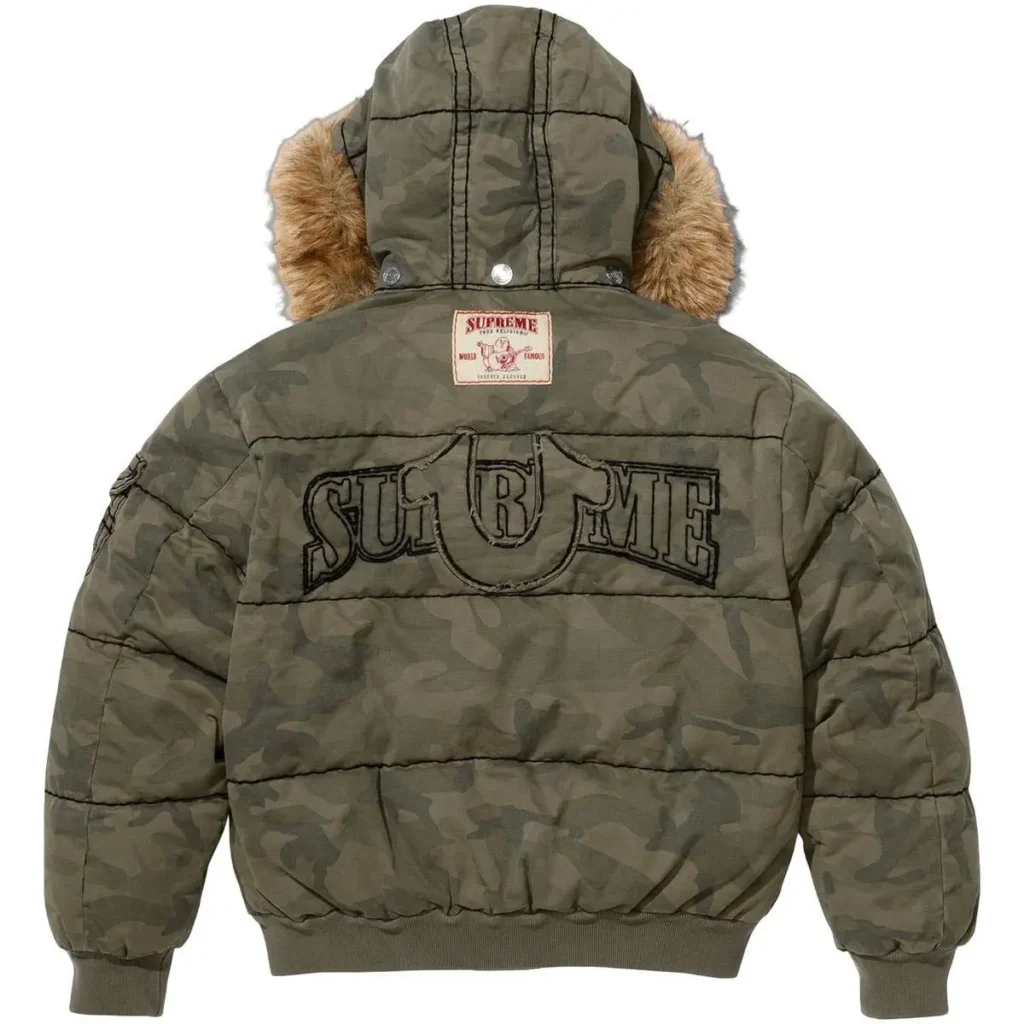 A camo Supreme × True Religion puffer jacket viewed from the back, featuring an oversized black embroidered Supreme horseshoe logo, a faux-fur trimmed hood, and a co-branded leather patch at the upper back