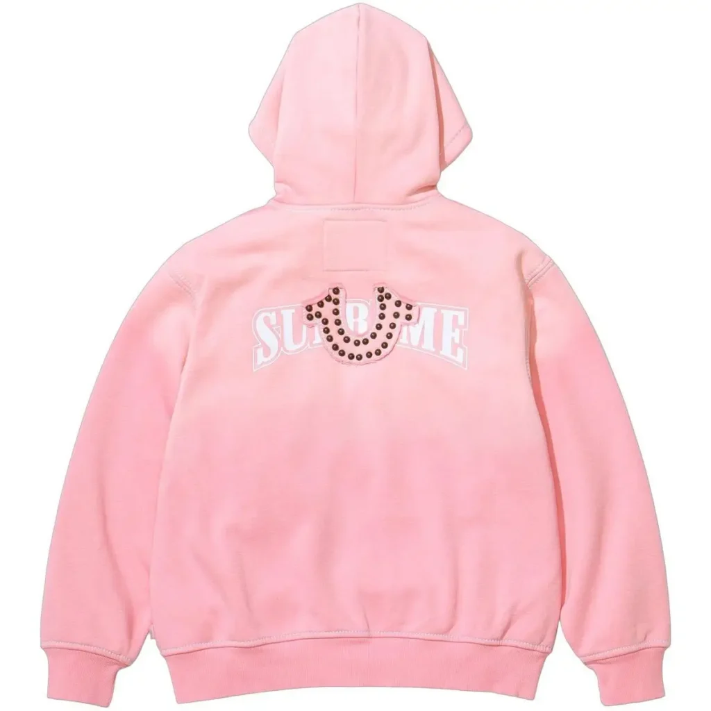 A pink Supreme × True Religion hooded sweatshirt shown from the back, featuring a white arched Supreme logo and a studded True Religion horseshoe emblem centered above it