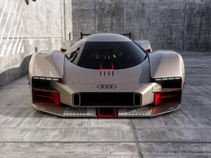 Front view of the Audi 20quattro Vision GT concept car showcasing its aggressive aerodynamic design, wide stance, illuminated Audi rings, and sharp LED headlight strips, with red aerodynamic accents and angular air intakes set against a concrete racing environment.