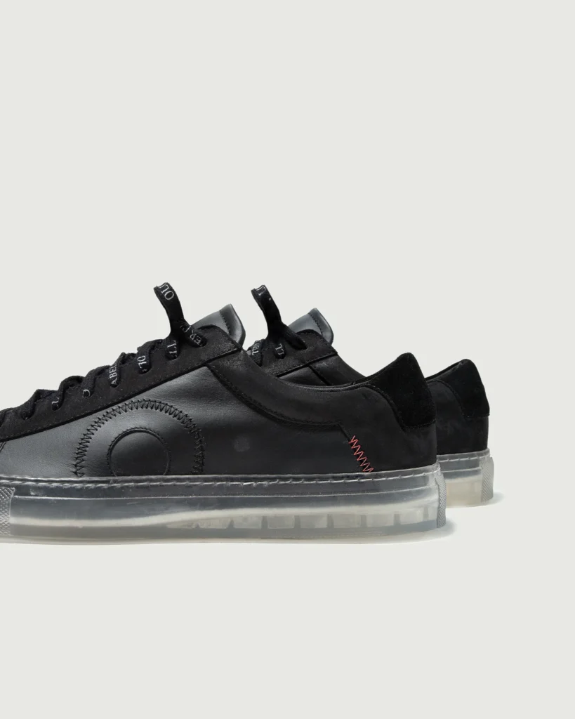 Side view of the Oliver Cabell Low 1 “Black Ghost” sneakers showing their matte black leather upper, circular stitched detailing, suede heel panels, and translucent rubber sole for a sleek, modern finish.