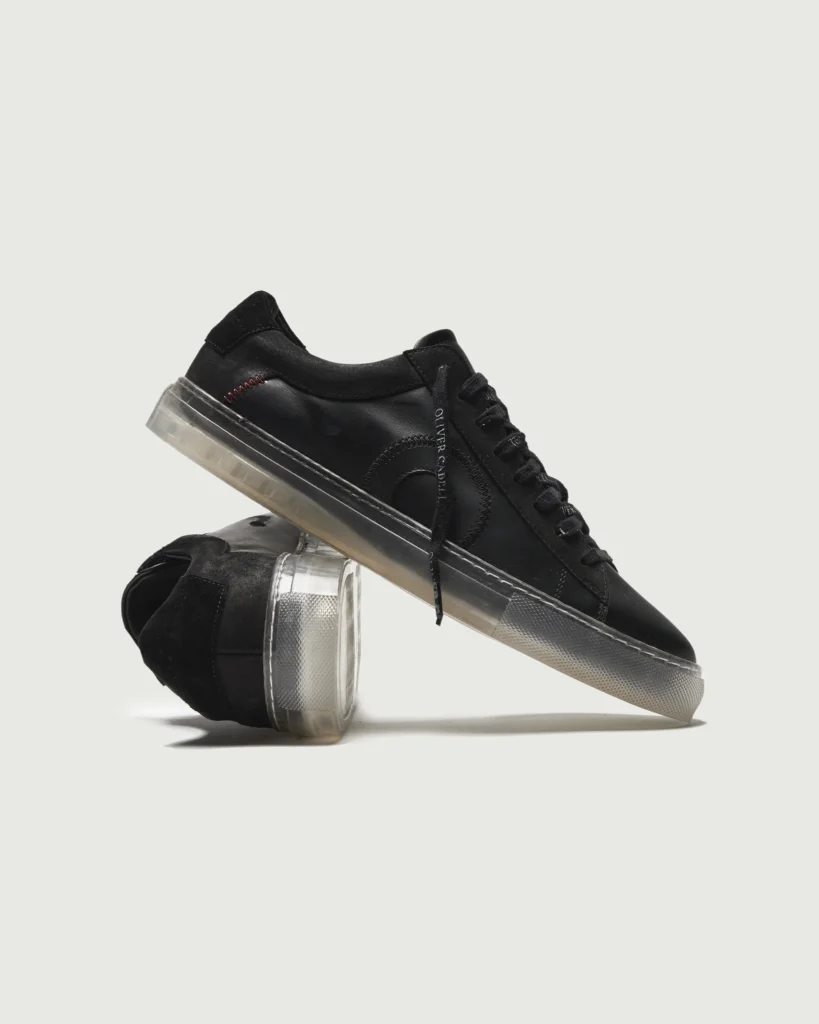 Two Oliver Cabell Low 1 “Black Ghost” shoes displayed in a stacked composition, highlighting the matte black leather finish, tonal laces, and translucent sole that blends modern design with orthopedic-inspired craftsmanship.