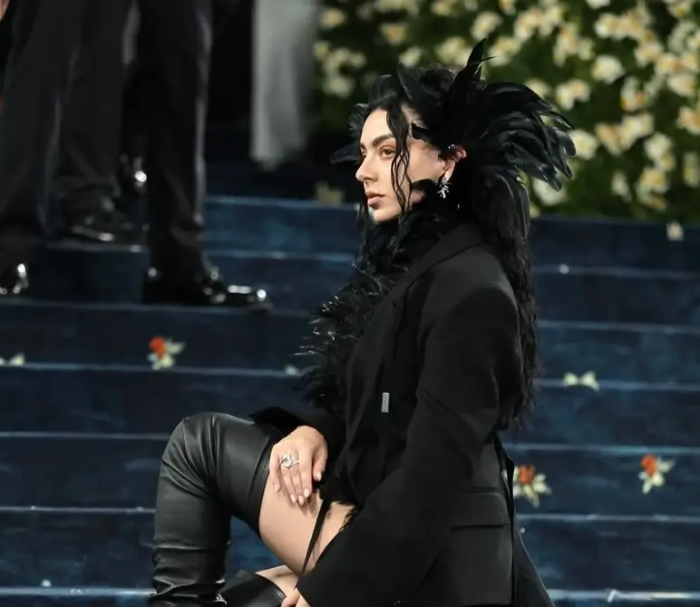 Charli xcx avant-garde black outfit sits on a staircase, wearing a dramatic feathered headpiece, long black coat, and thigh-high leather boots, with soft curls framing their face against a backdrop of blue carpeted steps and floral decorations