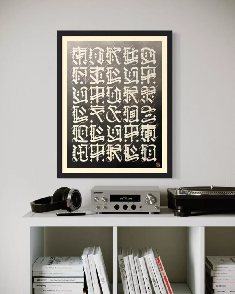 A framed artwork featuring dense cream-colored calligraphic glyphs on a black background hangs on a light grey wall. Below it sits a clean white shelf holding a pair of black over-ear headphones, a silver stereo receiver, and a black turntable. The lower cubbies contain neatly arranged books and magazines, creating a modern, minimalist home audio and art display