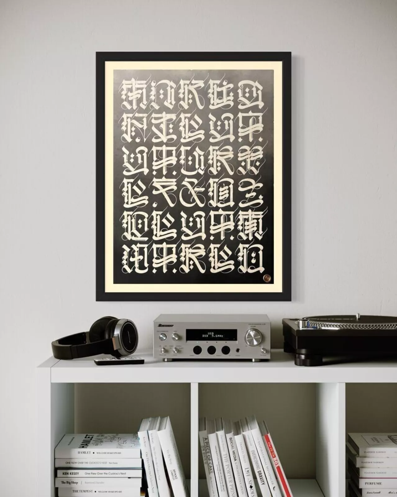 A framed artwork featuring dense cream-colored calligraphic glyphs on a black background hangs on a light grey wall. Below it sits a clean white shelf holding a pair of black over-ear headphones, a silver stereo receiver, and a black turntable. The lower cubbies contain neatly arranged books and magazines, creating a modern, minimalist home audio and art display