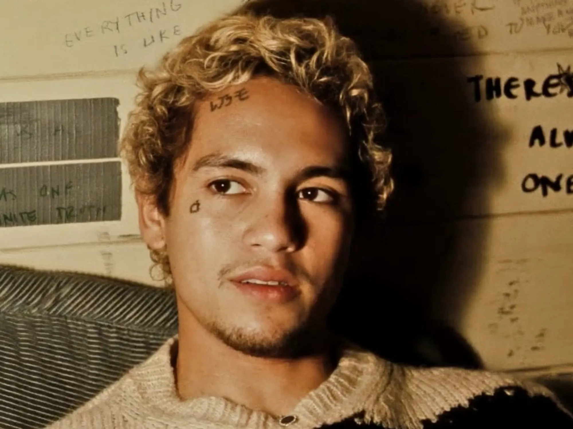 Dominic Fike with short curly blond hair sits in warm, low lighting, wearing a textured sweater, with small face tattoos visible as he looks slightly to the side against a wall covered in handwritten notes