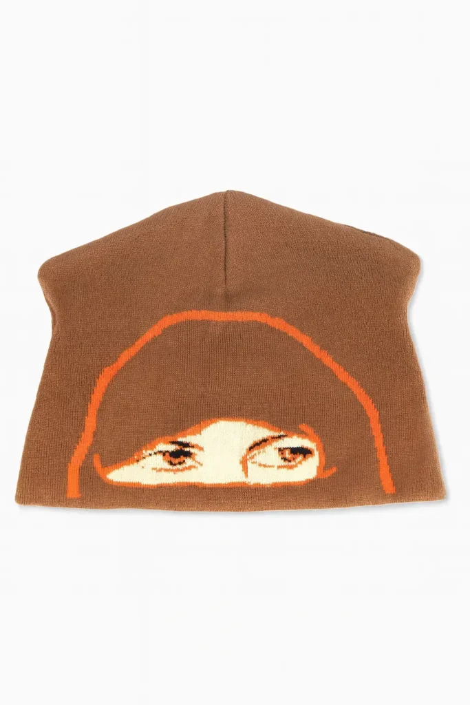 Brown knit skully beanie displayed on a white background, featuring an orange graphic outline of a hood framing a pair of illustrated eyes woven across the front