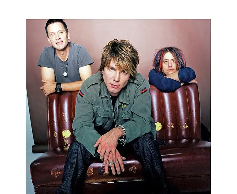 A portrait-style photo of three members of the Goo Goo Dolls sitting on a worn brown leather couch. The central figure sits forward with hands clasped, wearing a green military-style jacket and layered rings, with blonde tousled hair framing his face. To the left, another band member leans casually on the couch, smiling, wearing a grey T-shirt and pendant necklace. On the right, a third member with multicolored dreadlocks rests his arms on the couch back, looking toward the camera. The backdrop is a soft, neutral studio background
