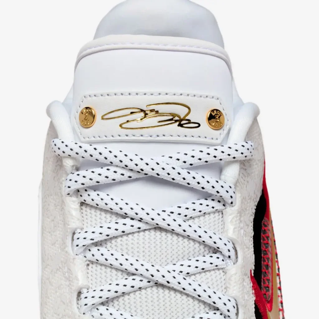 Close-up of the Nike LeBron 23 “Grand Opening” tongue, showing LeBron James’ gold-foil signature plate secured with two metallic gold rivets above white laces with black speckled detailing.