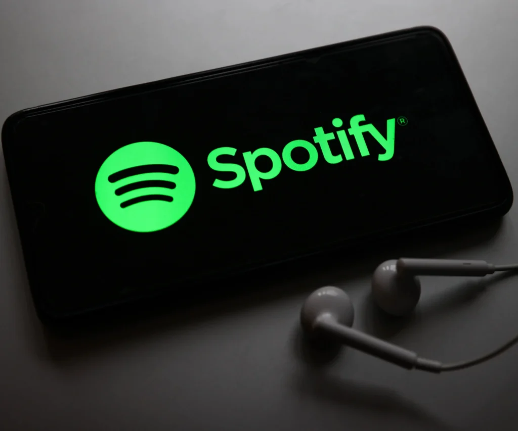 A smartphone laying on a flat surface displays the bright green Spotify logo on its screen, with a pair of white wired earbuds placed beside it