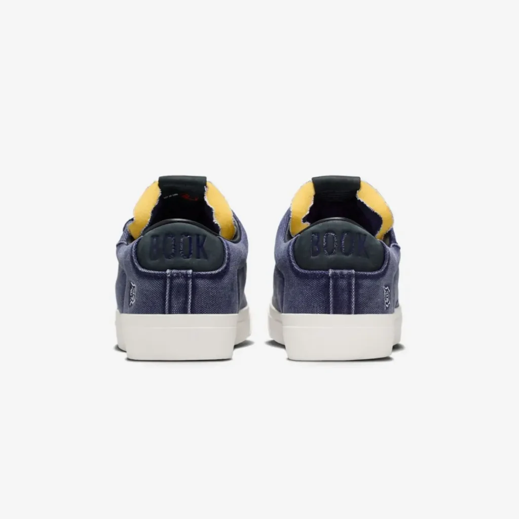 Rear view of a pair of navy blue low-top sneakers featuring embossed “BOOK” branding on the heel tabs, exposed yellow foam detailing, and white midsoles