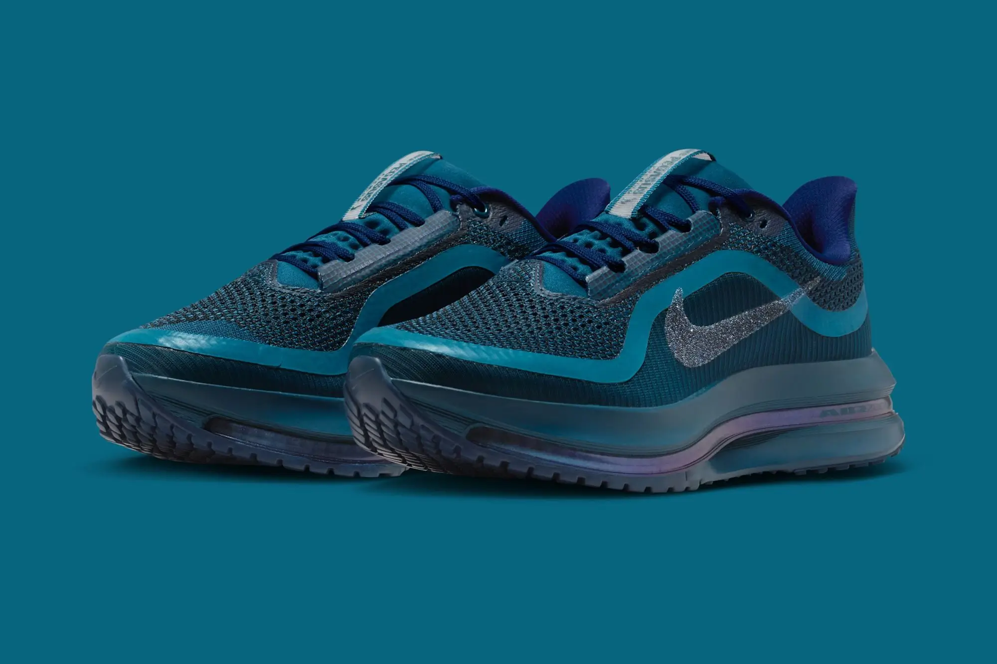 A pair of Swarovski × Nike Pegasus Premium SE WMNS “Blue Force” sneakers featuring a deep teal-blue mesh upper, tonal overlays, and a glittering Swarovski crystal-encrusted Nike Swoosh. The shoes rest on a sculpted midsole with a translucent Air Zoom cushioning unit, presented against a matching blue backgroun