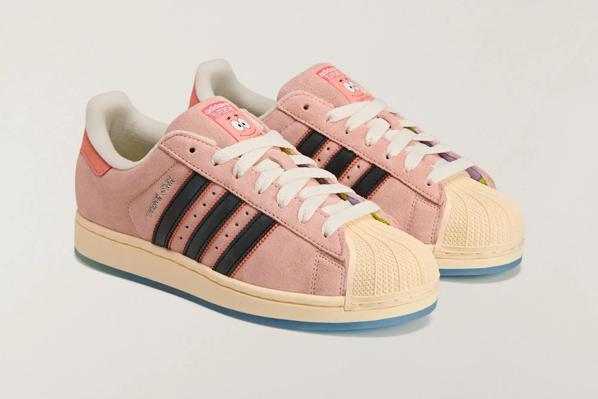 A pair of SpongeBob SquarePants x adidas Superstar “Patrick Star” sneakers featuring soft pink suede uppers, black Three Stripes with coral stitching, cream shell toes, white laces, and Patrick’s smiling face printed on the pink tongue labels, shown angled on a light neutral background