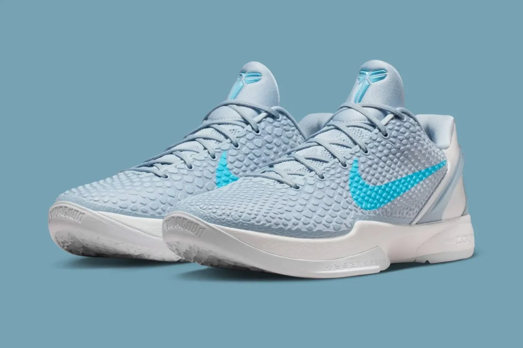 The Nike Kobe 6 Protro “Caitlin Clark” sneaker displayed in a three-quarter view, featuring a light icy-blue snakeskin-textured upper with a bright blue Nike Swoosh on the side. The shoe sits on a white midsole with subtle sculpting, while the tongue shows Kobe Bryant’s signature sheath logo in matching blue. The background is a solid light blue, highlighting the sneaker’s cool, monochromatic palette