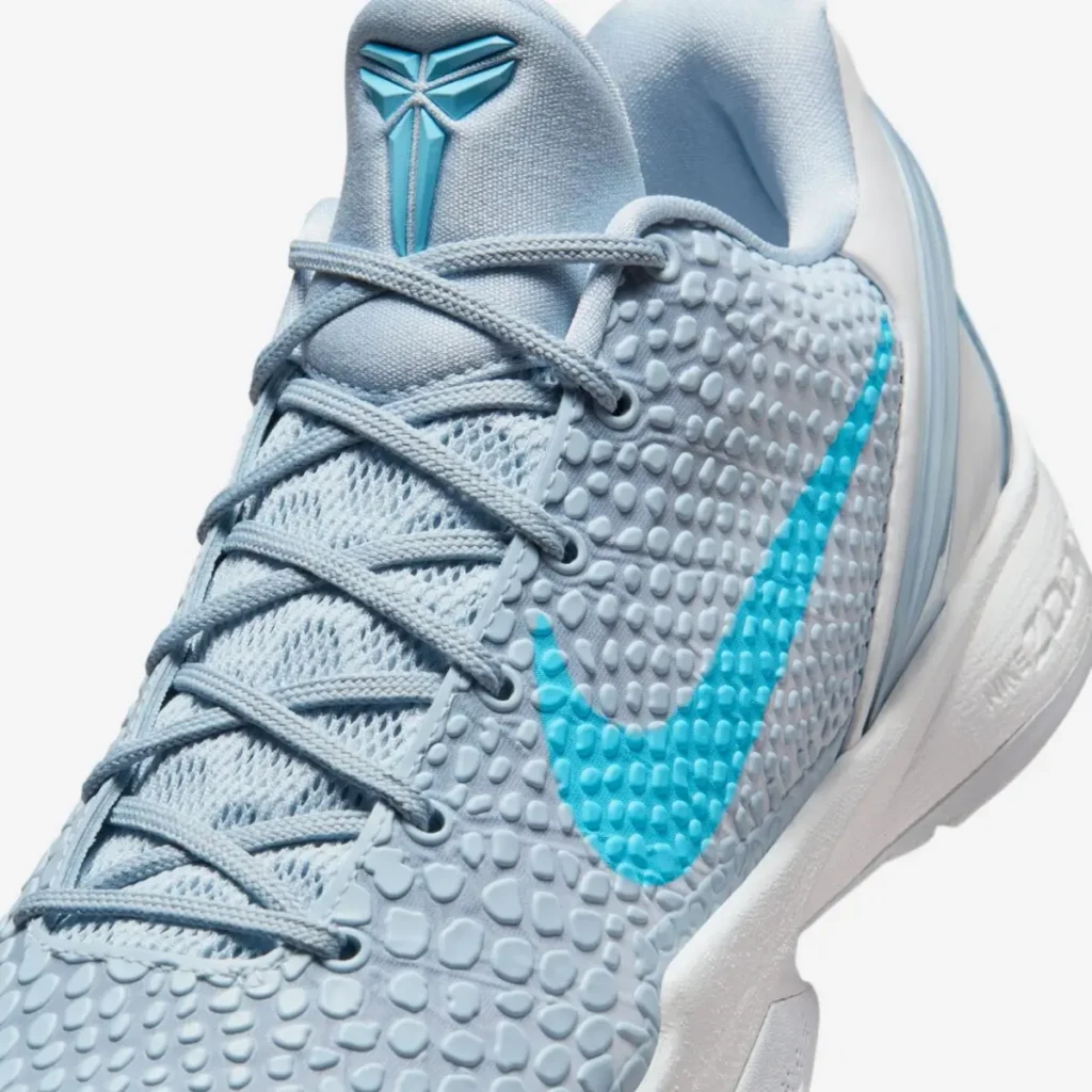 A detailed close-up of the Nike Kobe 6 Protro “Caitlin Clark,” showing the light icy-blue snakeskin-textured upper, matching woven laces, and bright blue Nike Swoosh. The padded tongue features Kobe Bryant’s sheath logo in a glossy blue finish, while the white midsole and sculpted heel counter appear along the edge of the frame