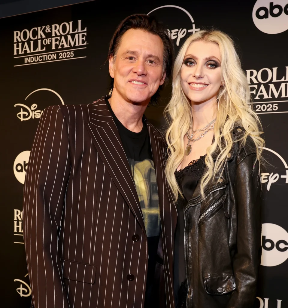 Jim Carrie and Taylor Momsen standing together on a black carpet at a Rock & Roll Hall of Fame event, smiling toward the camera. One wears a dark pinstriped blazer while the other wears a black leather jacket and layered silver necklaces