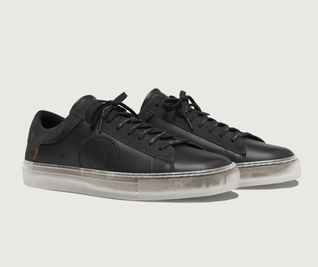 A pair of Oliver Cabell Low 1 “Black Ghost” shoes made from matte black Italian leather with tonal stitching, suede heel panels, and a translucent rubber sole, showcasing the brand’s minimalist design and orthopedic-inspired construction.