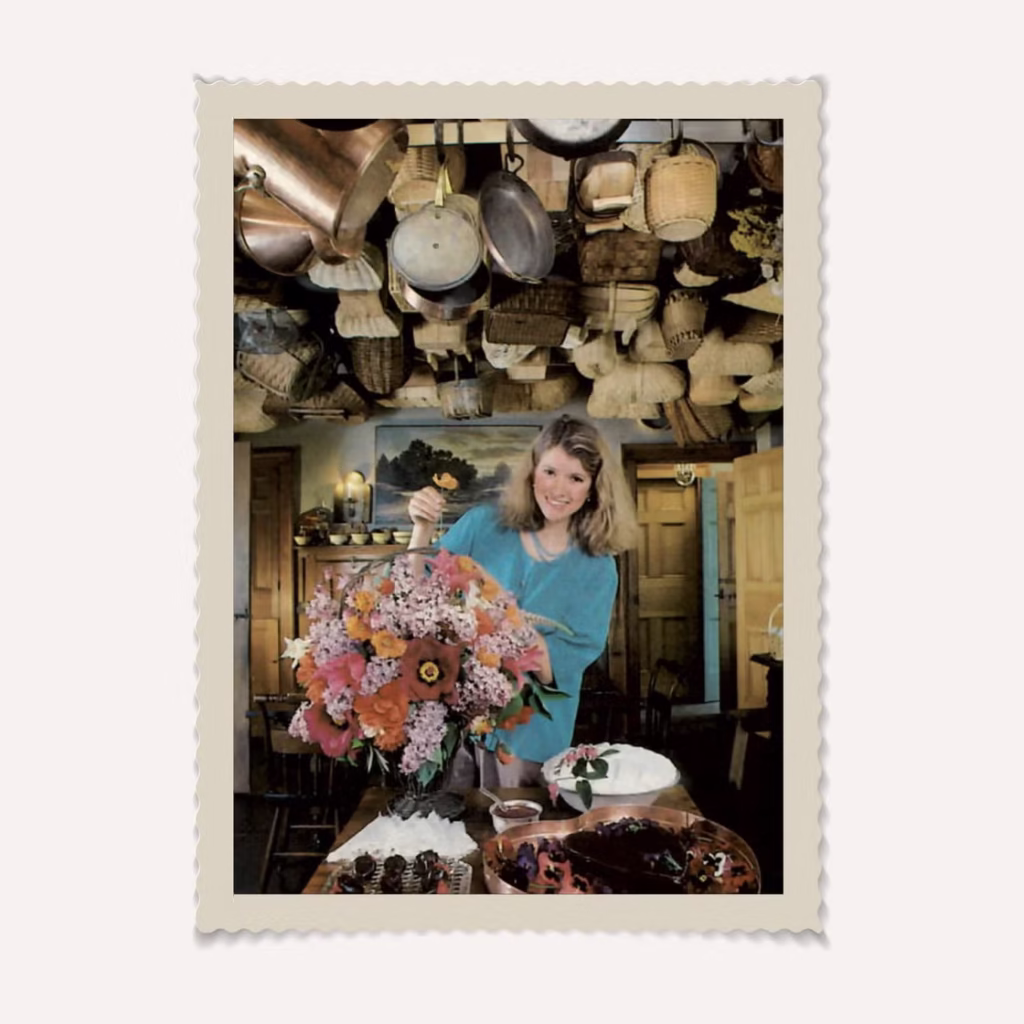 Martha Stewart seated in a warmly lit kitchen, surrounded by table settings and floral arrangements that echo the aesthetic of her 1982 book Entertaining, symbolizing the timeless charm of her early lifestyle vision ahead of its 2025 facsimile reissue