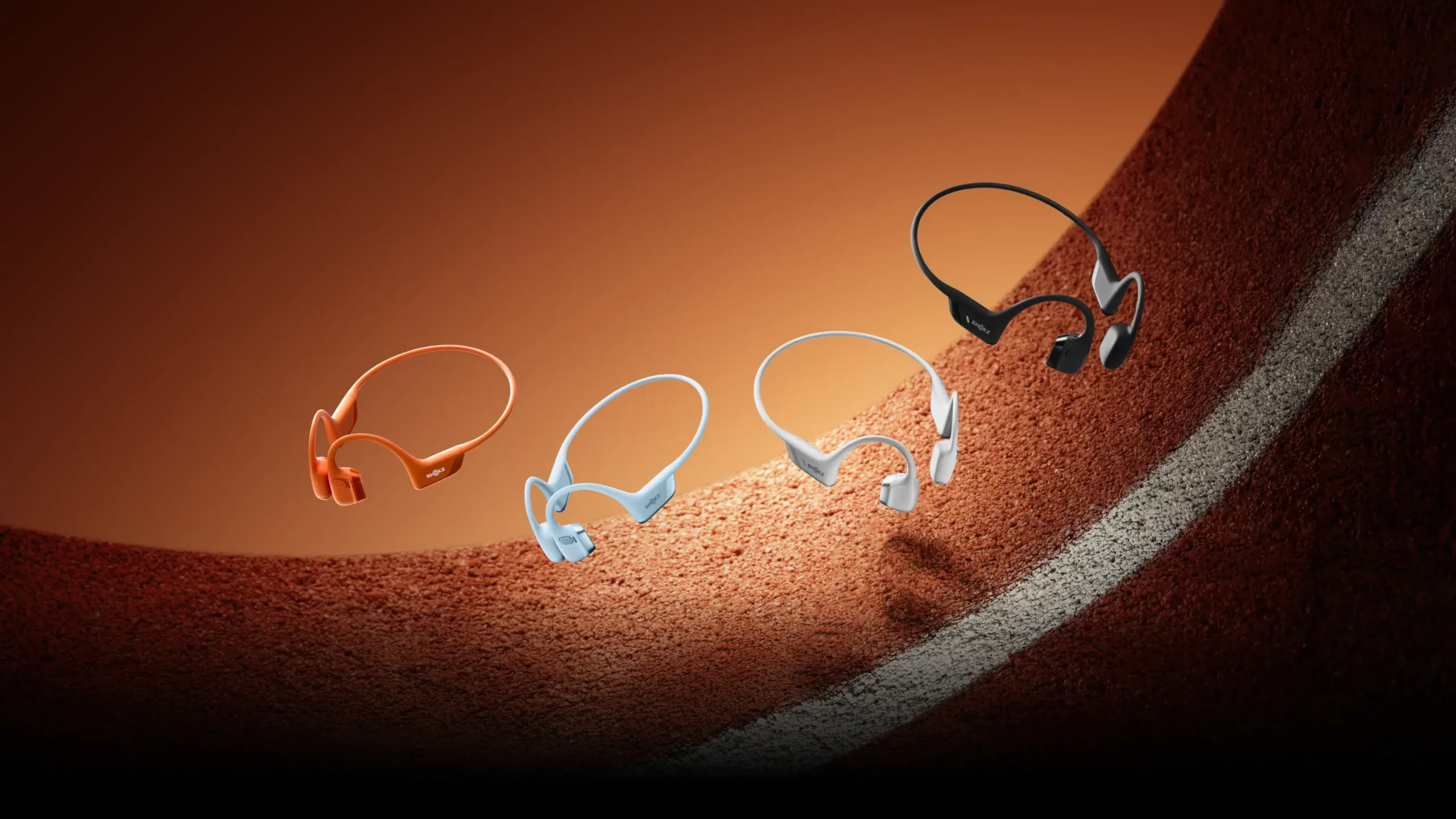 Four Shokz OpenRun Pro 2 bone-conduction sports headphones in orange, sky blue, white, and black appear suspended above a curved running-track surface under warm studio lighting, symbolizing speed, endurance, and motion.