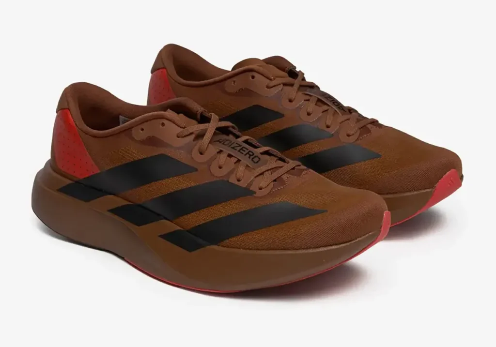 Close-up of the Pharrell Williams x adidas Adizero EVO SL “Preloved Brown” sneakers showing the deep brown mesh upper with matte black three-stripes, red heel panel, and sculpted midsole—captured on a clean white background highlighting the shoe’s streamlined silhouette and earthy tones