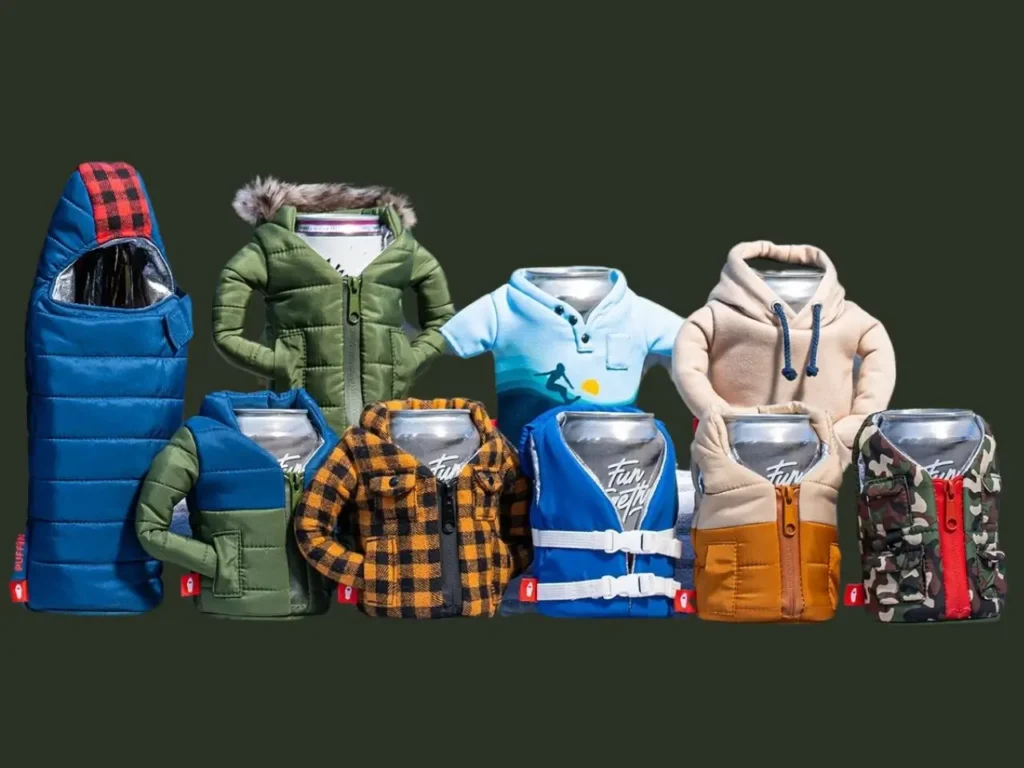 A lineup of Puffin Drinkware holders styled as miniature outerwear, including puffer jackets, flannel shirts, fleece hoodies, vests, life jackets, and a sleeping-bag–style cover. The display features a variety of colors and patterns—greens, blues, camo, plaid, and pastels—with each insulated holder fitted around a beverage can, creating a playful collection of tiny winter-themed outfits