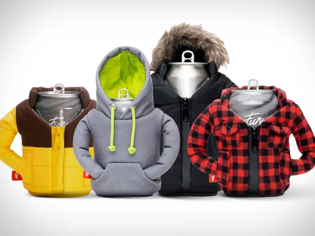A lineup of beverage cans dressed in miniature Puffin Drinkware outfits, including a yellow puffer jacket, a grey hoodie with neon lining, a black parka with faux fur hood, and a red-and-black plaid flannel jacket. Each insulated holder mimics real winter outerwear with zippers, pockets, and detailed stitching