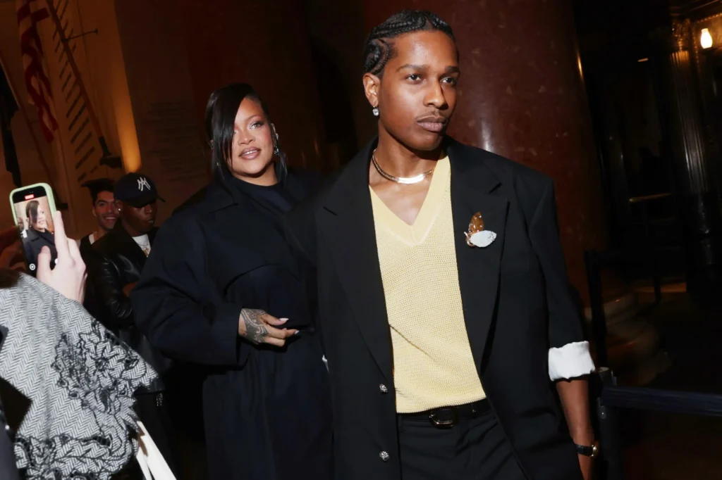Rihanna and A$AP Rocky pose together at the 2025 CFDA Fashion Awards in New York; Rihanna wears a white trench and matching trousers, while A$AP Rocky stands beside her in a black suit and yellow knit shirt, holding his Fashion Icon Award