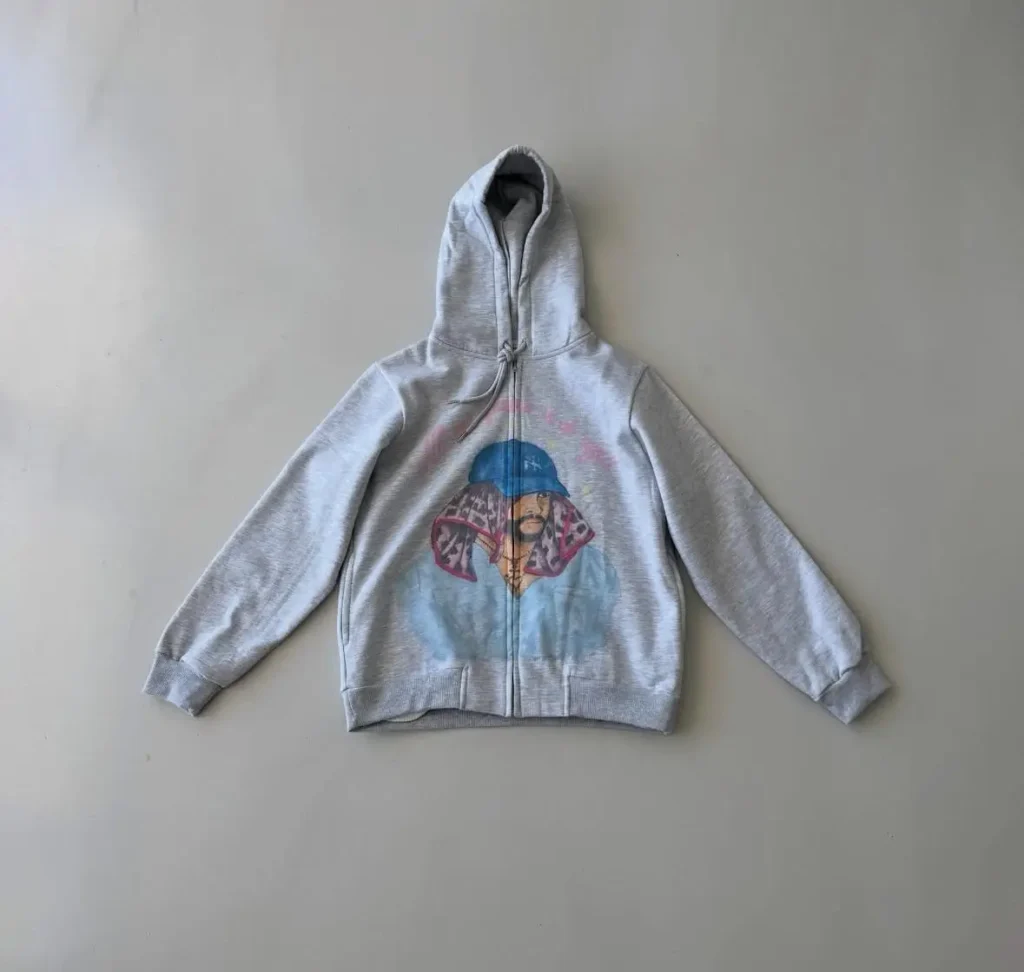 Light grey full-zip hoodie featuring a colorful illustrated portrait of Bad Bunny wearing a blue hat and patterned scarf, with the phrase “No me quiero ir de aquí” printed above in pink lettering — part of the official Bad Bunny × FRSH Company Puerto Rico residency merchandise collection