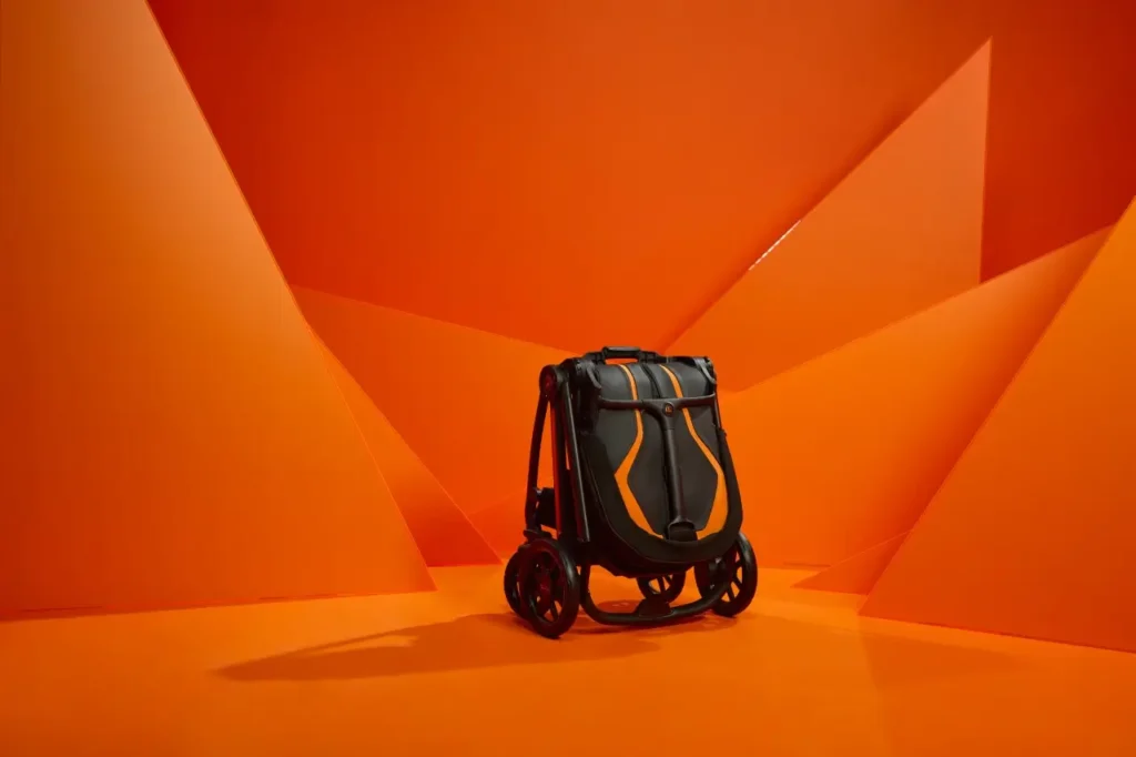 The Silver Cross × Lamborghini stroller shown in its folded, compact form, featuring black and orange panels with aerodynamic lines reminiscent of a Lamborghini supercar. The stroller stands upright against a bold, geometric orange backdrop that enhances its sporty, high-performance aesthetic