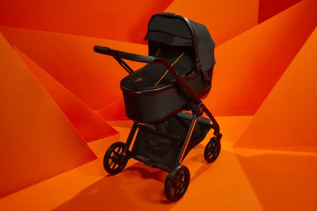 Black Silver Cross × Lamborghini stroller with angular matte-black frame, orange accent piping, and Lamborghini crest on the bassinet, photographed against a geometric bright-orange backdrop that echoes the brand’s signature supercar palette