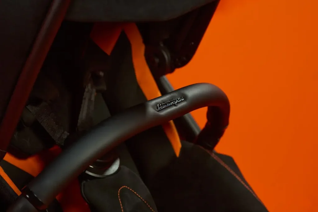 Close-up of the Silver Cross × Lamborghini stroller’s matte-black handlebar, featuring an embossed Lamborghini logo. The surrounding fabrics show black upholstery with orange contrast stitching, set against a vivid orange backdrop that highlights the stroller’s luxury automotive-inspired detailing