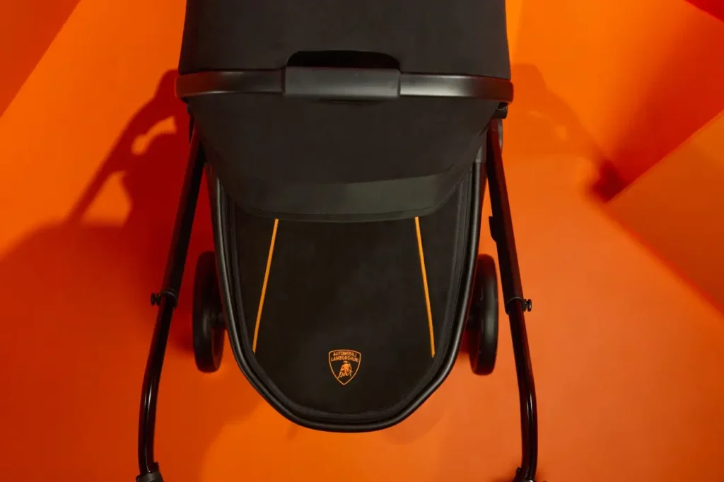Top-down view of the Silver Cross × Lamborghini stroller’s footmuff panel, featuring black suede-like fabric, two vertical orange accent stripes, and the Automobili Lamborghini crest centered near the bottom. The stroller sits against a vibrant orange geometric backdrop matching the brand’s signature color palette