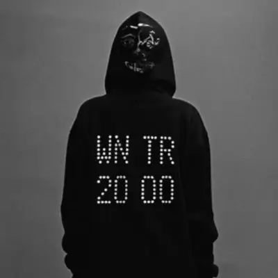 IDK wearing a black hoodie stands with their back to the camera against a gray background. The back of the hoodie displays the text “WN TR 20 00” in a dotted, digital-style font. The hood is up, partially revealing a stylized skull graphic printed on the back of the hood