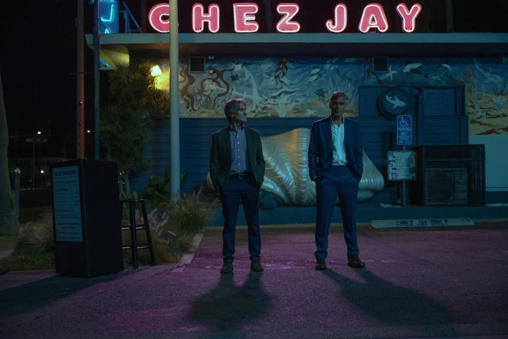 Two men stand outside the neon-lit Chez Jay restaurant at night, illuminated by moody blue and pink lighting, creating a cinematic atmosphere suggestive of a tense or reflective moment