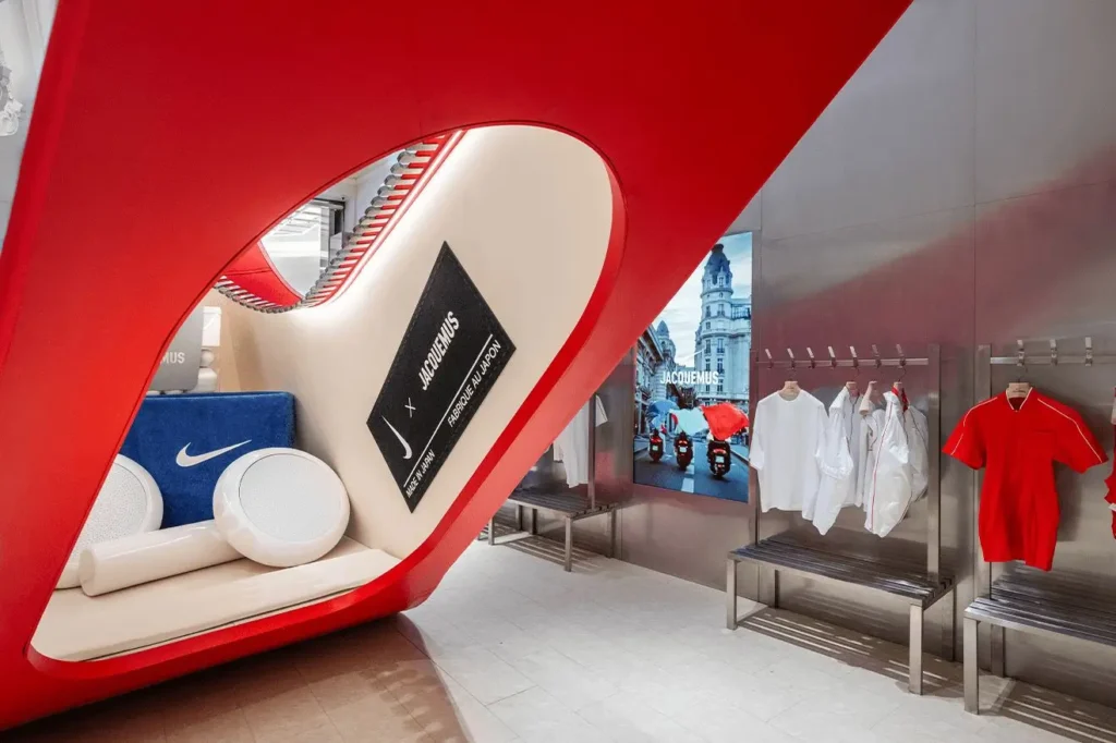 Interior view of the Nike × Jacquemus installation at Selfridges, showing the underside of a large red sculptural swoosh with beige cushioning, oversized white gym-inspired props, a blue Nike-branded block, and a display of white and red Jacquemus apparel against metallic walls