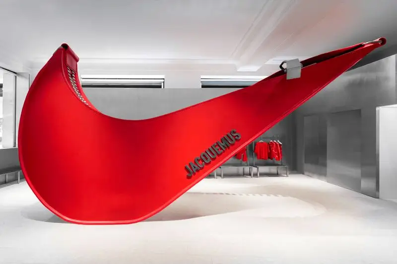 A large sculptural red Nike swoosh installation with “Jacquemus” lettering displayed inside Selfridges, set in a minimalist silver-toned retail space with coordinated red apparel hanging in the background