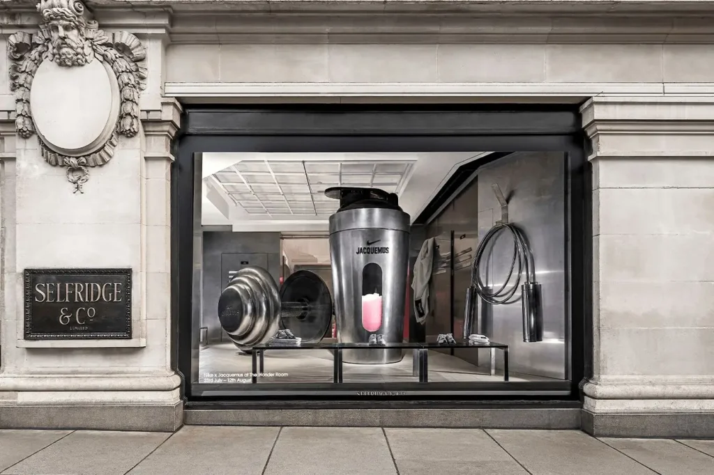 Exterior view of the Selfridges window display featuring the Nike × Jacquemus installation, showcasing oversized metallic gym objects including a giant shaker bottle, weight plates, and a coiled jump rope inside a sleek industrial-style interior
