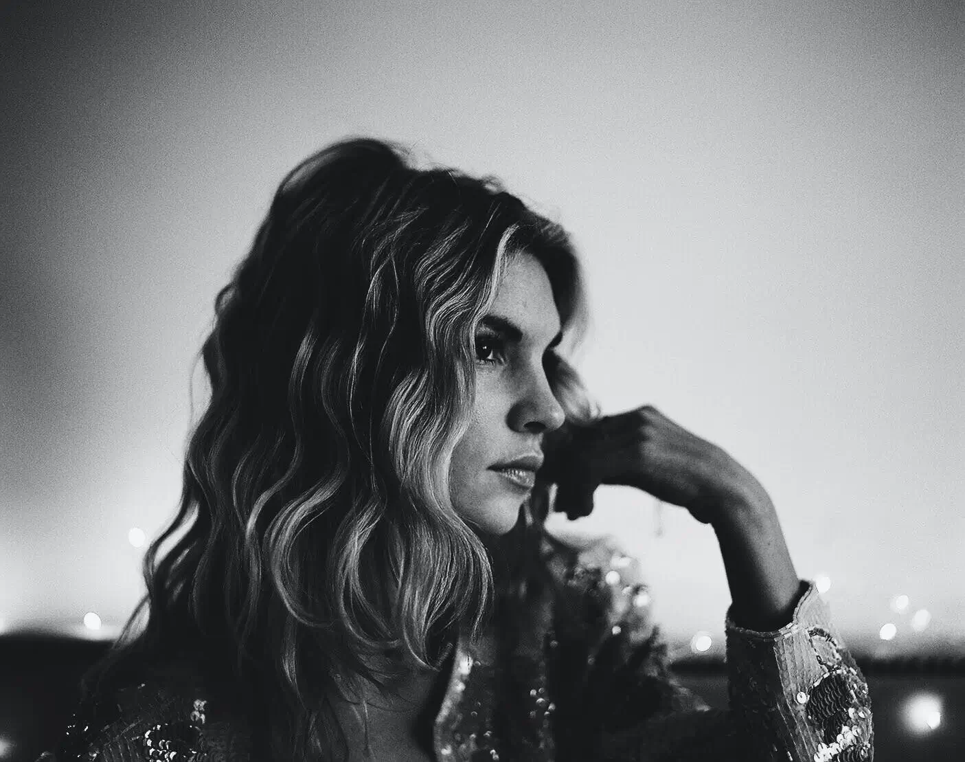 A black-and-white portrait of a woman with wavy, shoulder-length hair, seated in profile with her hand resting against her face, wearing a sequined jacket as soft lights glow faintly in the blurred background