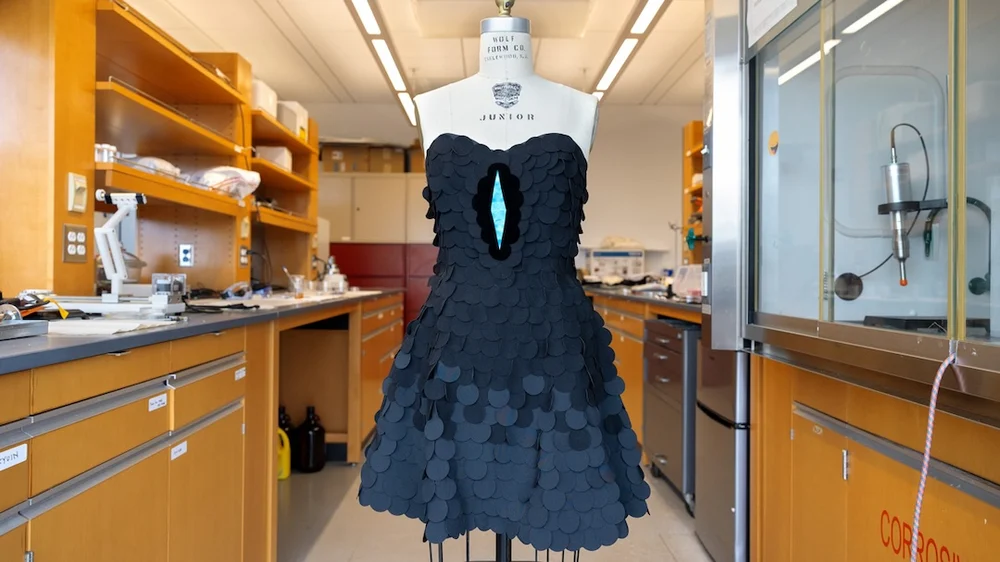 A dress form in a Cornell University laboratory displaying a prototype dress made from ultrablack fabric scales, with a central iridescent blue slit inspired by a bird-of-paradise display. The surrounding workspace shows lab equipment, shelves, and scientific tools, highlighting the experimental context of the textile