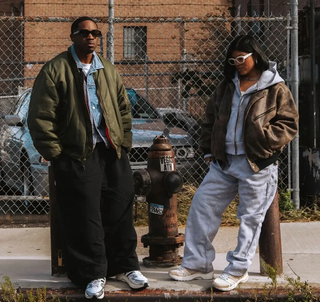Two people wearing street-style outfits stand beside an old fire hydrant in an urban setting, with both dressed in oversized outerwear and retro-tech adidas running sneakers. The background features a chain-link fence, parked cars, and brick buildings, creating a gritty NYC-inspired atmosphere