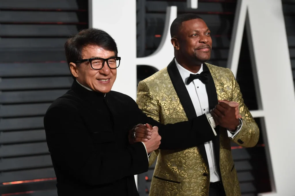 Chris Tucker and Jackie Chan pose together at a formal event, smiling and clasping hands in a friendly gesture. The man on the left wears black-framed glasses and a sleek black mandarin-style jacket, while the man on the right wears a gold embroidered tuxedo jacket with black lapels and a bow tie. The backdrop features a modern black-and-white design with horizontal paneling
