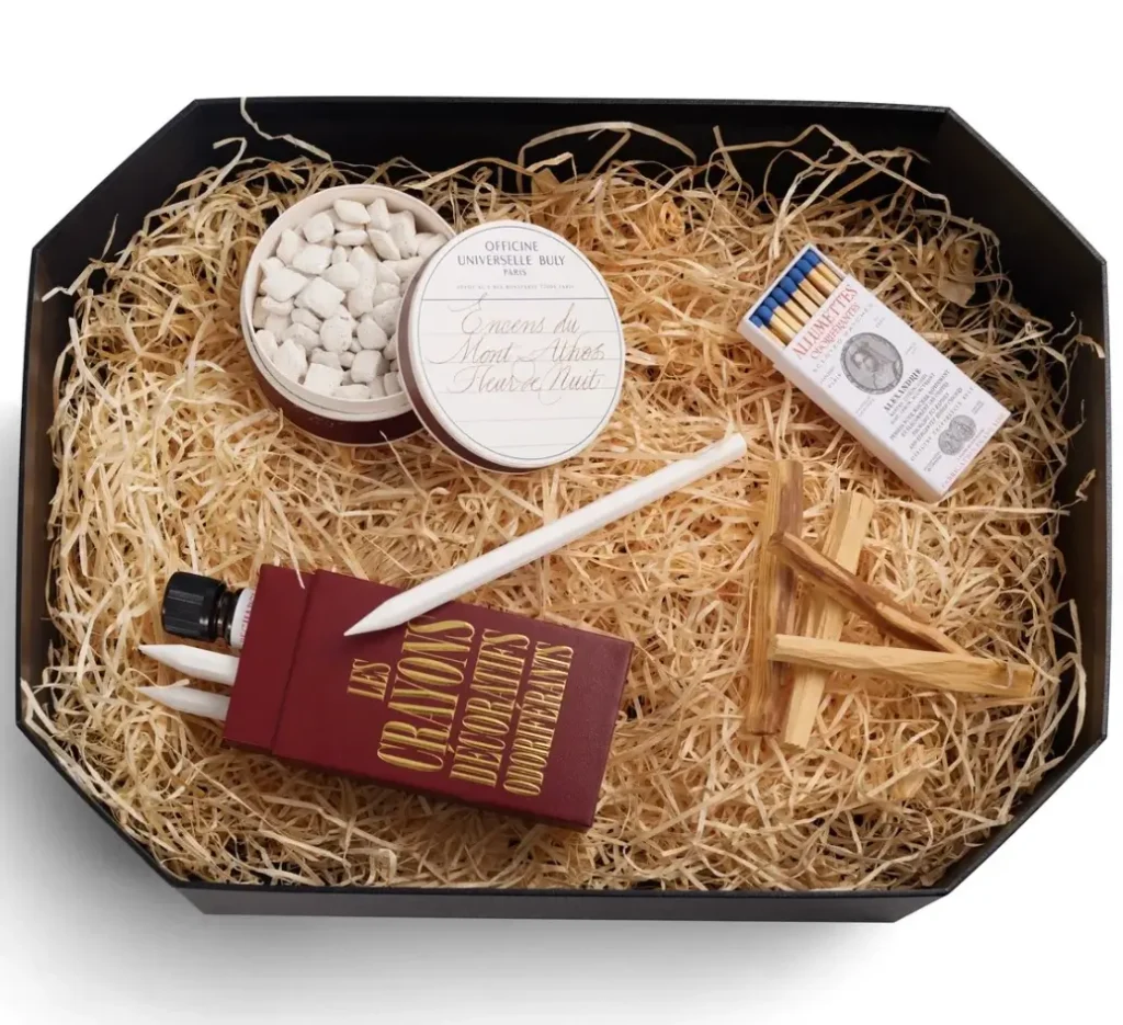 Octagonal gift box filled with shredded straw, displaying Officine Universelle Buly’s scented interior set including a tin of Mount Athos incense pellets, a box of long scented matches, Palo Santo wood sticks, and a burgundy case of decorative scented pencils with diffuser oil